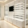 Modern Glossy White Wall Eyewear Display Wall | LED Lit Adjustable Glass Shelving Optical Display Unit With Central Digital Screen & Multi-Drawer Base Cabinets For Contemporary Optical Shop Fit-Out
