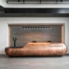 Sculptural capsule‑shaped aged copper reception desk with rounded bulbous ends and integrated top LED strip, large luxury lobby front desk Lexio Display for high‑end commercial fit‑out