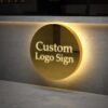 Custom round backlit metal logo sign on textured brown lobby wall, warm halo backlight