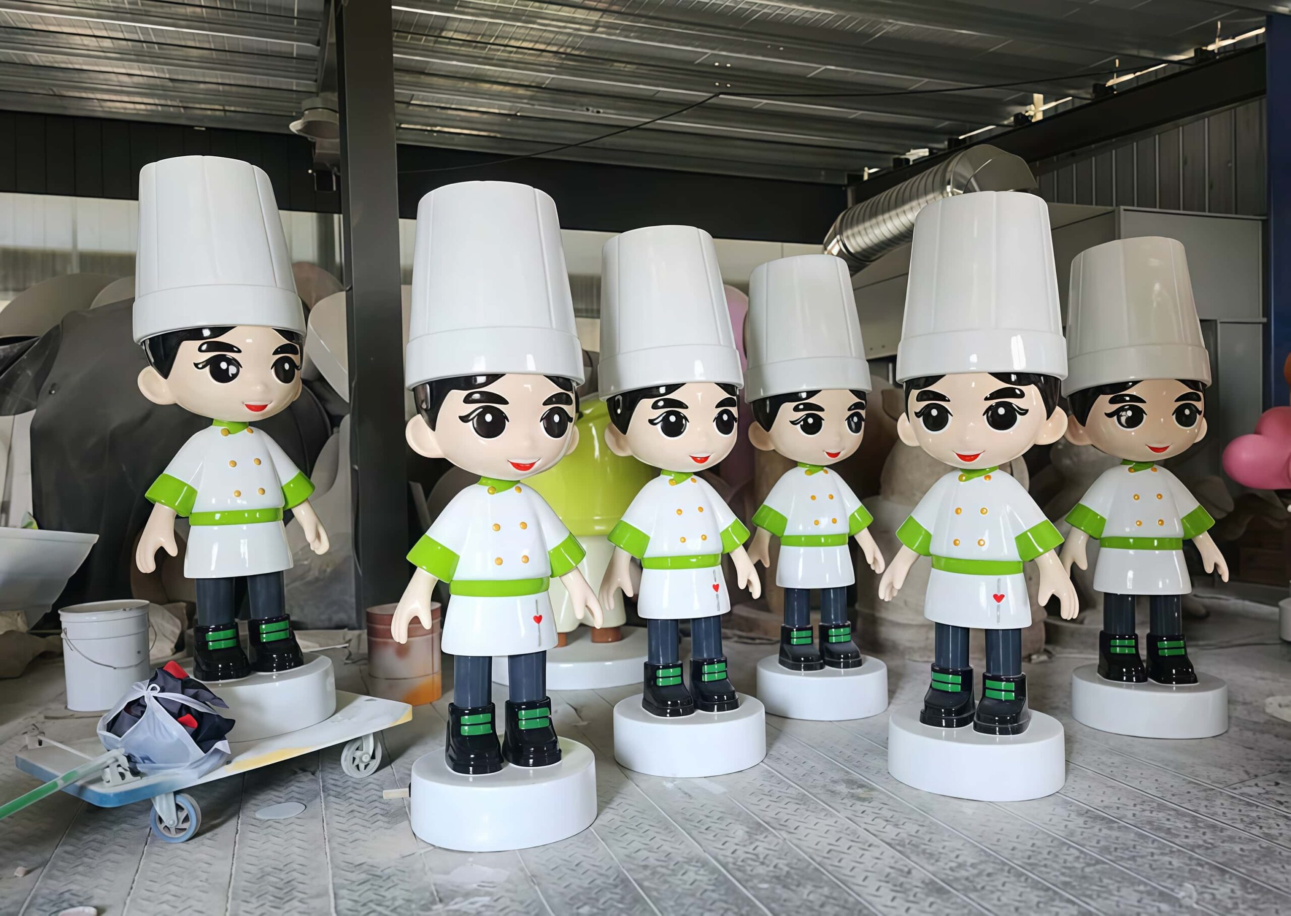 Factory workshop photo of batch cartoon chef fiberglass mascot sculptures, chibi chef characters with tall white chef hats and green trimmed chef coats